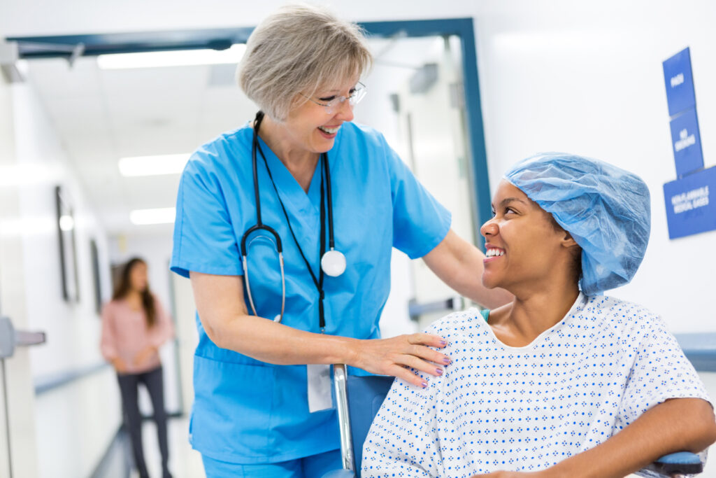 Senior adult Caucasian female nurse with grey hair is pushing patient in wheelchair. She is taking patient to recovery after outpatient surgical procedure. Patient is young adult African American woman, who is wearing a hospital gown and surgical cap. Women are smiling while talking to each other.