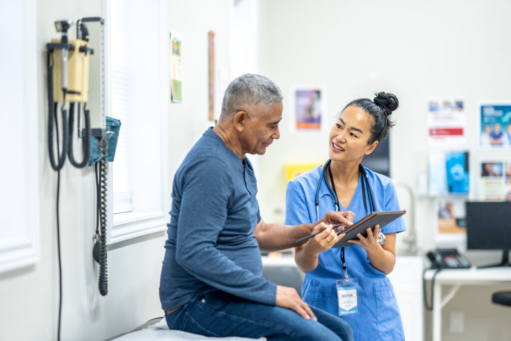 A senior gentleman of Hispanic decent, sits up on an exam table during a routine check-up at the doctors.  A nurse, who is wearing blue scrubs, is standing in front of the gentleman as she reviews some recent test results with the patient on a tablet.